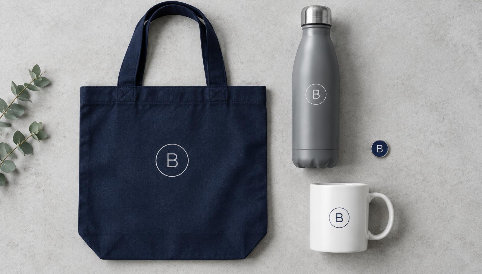 Flat-lay of branded navy tote bag, grey water bottle, white mug and enamel pin